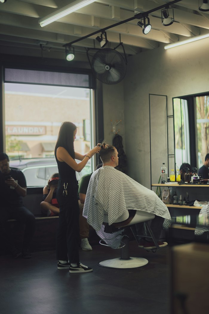 The barber standing behind a caped client in a barber chair, scissors in hand, in a bright shop